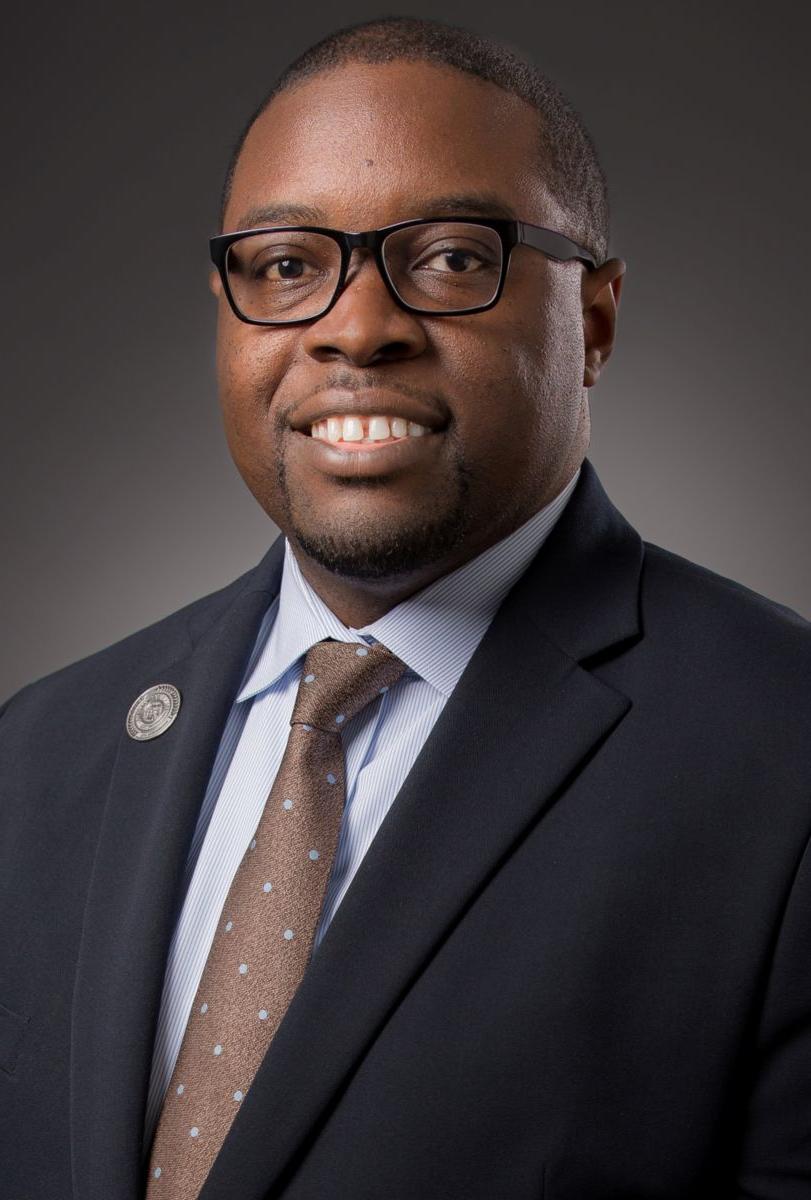 Dr. Jermaine Whirl, an African American male wearing a grey suit jacket, a light blue collared shirt, black rimmed glasses, and a cerulean blue tie. A gold pin is on his suit jacket lapel.